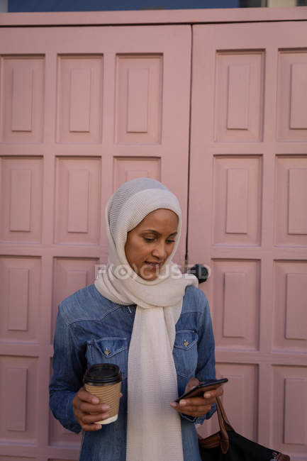 Вид спереду красива змішаної раси жінка використовує мобільний телефон і тримає каву, стоячи перед дверима на вулиці — стокове фото