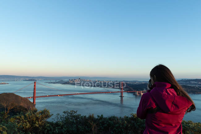 Vista trasera de la mujer tomando una foto de un puente en un día soleado - foto de stock
