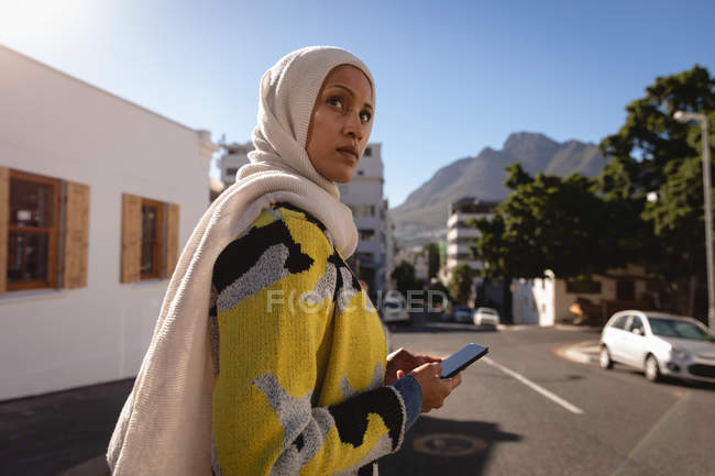 Side view of a mixed-race woman using mobile phone while walking over the middle of street on a sunny day — Stock Photo