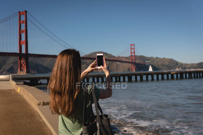 Vista posteriore della donna che cattura l'immagine con il telefono cellulare del ponte sospeso — Foto stock
