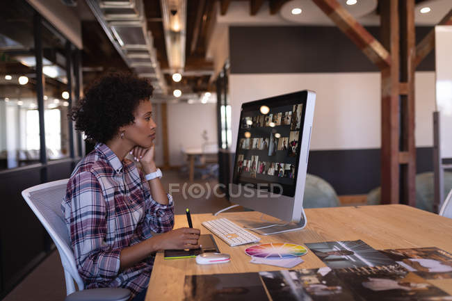 Vista lateral del hermoso diseñador gráfico femenino de raza mixta utilizando una tableta gráfica en el escritorio mientras mira su computadora - foto de stock