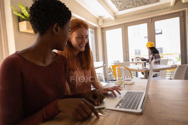 Vista lateral de jóvenes amigas de raza mixta que interactúan entre sí mientras usan el portátil en un café - foto de stock
