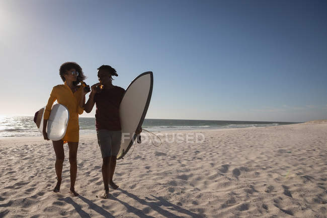 Vue de face du couple afro-américain buvant de la bière tout en tenant skateboard à la plage le jour ensoleillé — Photo de stock