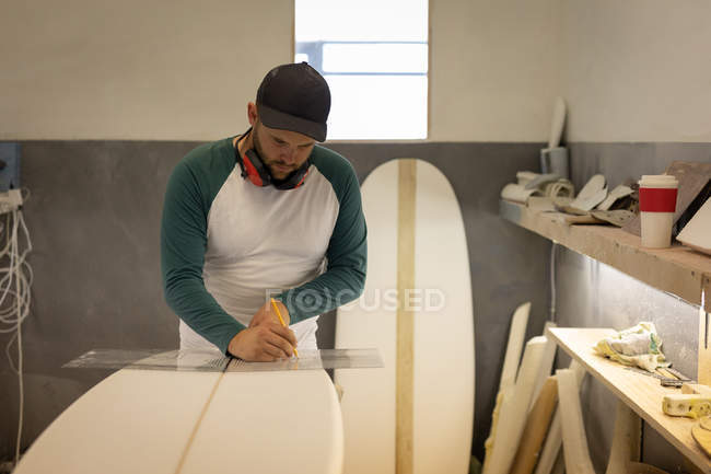 Vista frontal de hombre caucásico guapo con orejeras que miden con regla y lápiz una tabla de surf en el taller - foto de stock