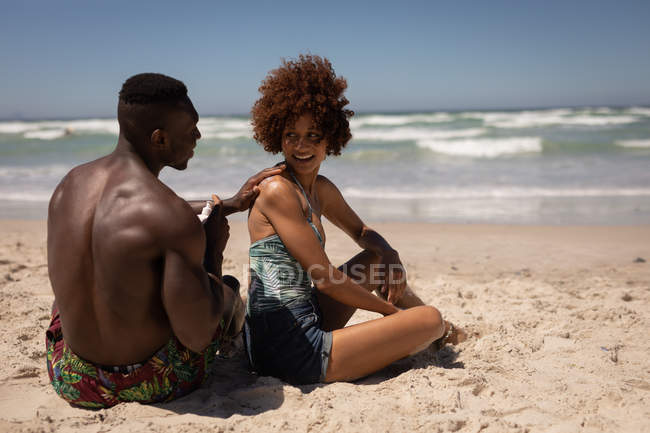 Vue arrière d'un homme afro-américain heureux appliquant une crème solaire sur le corps d'une femme métisse à la plage — Photo de stock