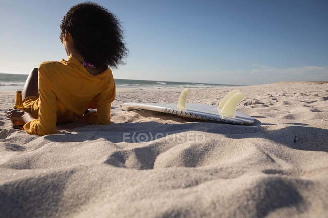 Vista trasera de la hermosa mujer afroamericana inclinada en la playa mientras toma cerveza en un día soleado - foto de stock