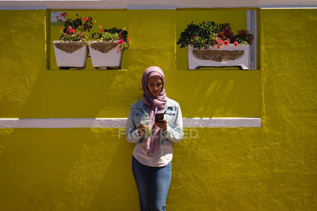 Vorderansicht der schönen Mischlingshündin, die an einem sonnigen Tag ihr Handy benutzt, während sie sich an die Wand lehnt — Stockfoto