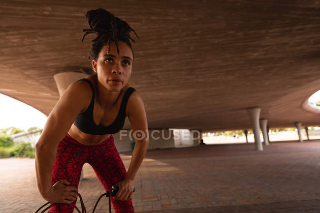 Vista frontal da jovem mulher de raça mista descansando depois de se exercitar sob uma ponte na cidade — Fotografia de Stock