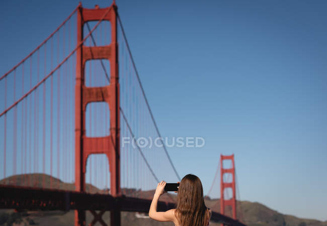 Visão traseira da mulher capturando imagem com telefone celular de ponte suspensa — Fotografia de Stock