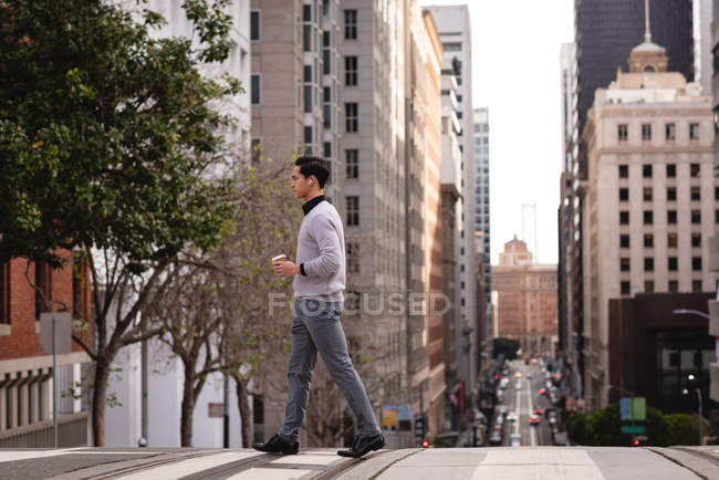 Vista lateral do homem asiático atravessando a rua enquanto toma café — Fotografia de Stock