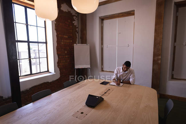 Вид спереду бізнесмен змішаної раси, що пише на щоденнику в творчому офісі на білій дошці на задньому плані — стокове фото