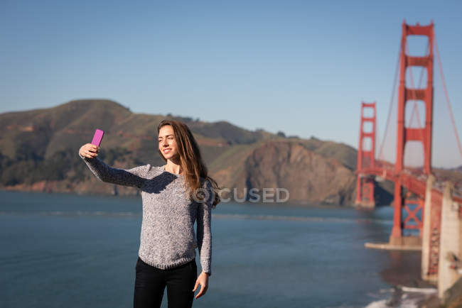 Front view of beautiful woman taking selfie in front of suspension bridge — Stock Photo