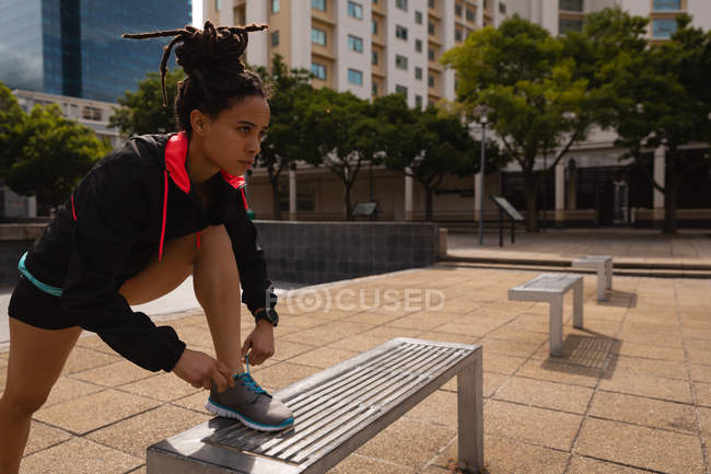 Vista laterale della giovane donna di razza mista che si lega i lacci delle scarpe su una panchina in città — Foto stock