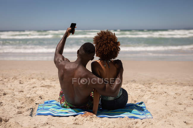 Vista trasera de feliz pareja multiétnica tomando selfie con teléfono móvil en la playa - foto de stock