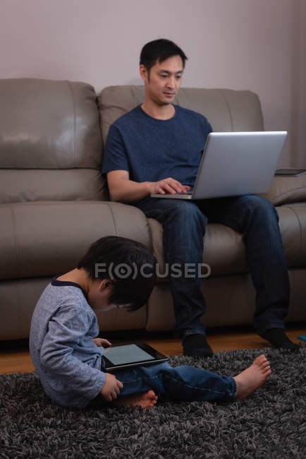 Vue de face du jeune père asiatique utilisant un ordinateur portable sur le canapé tandis que son fils utilisant une tablette numérique à la maison — Photo de stock