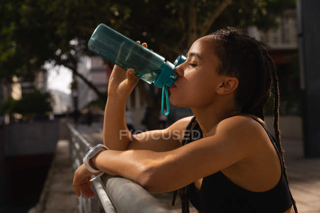 Vista lateral de la joven cansada Mujer de raza mixta bebiendo agua en la ciudad - foto de stock