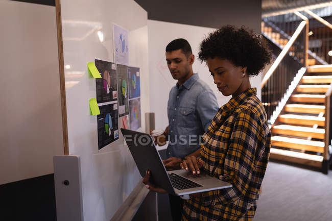 Vista lateral da jovem mulher de negócios de raça mista usando laptop enquanto homem de negócios de raça mista em pé ao lado do gráfico no escritório moderno — Fotografia de Stock