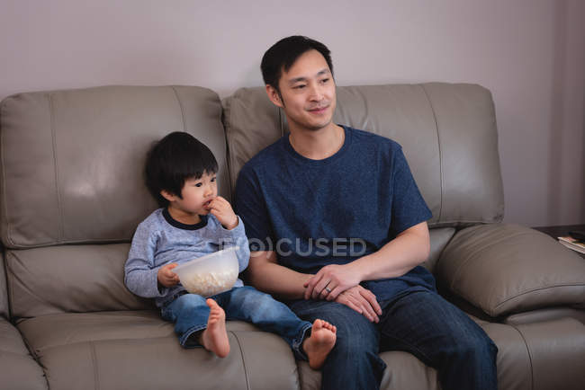 Вид спереди счастливого азиатского отца и сына, сидящих на диване и поедающих попкорн, смотря телевизор дома — стоковое фото