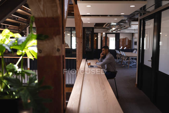 Vue latérale d'un homme d'affaires métis parlant sur un téléphone portable tout en utilisant un ordinateur portable dans un bureau moderne — Photo de stock