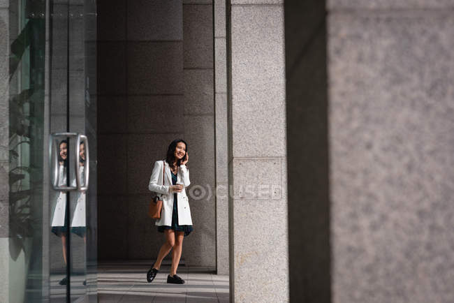 Vista lateral de la joven hermosa mujer asiática hablando por teléfono móvil mientras camina por el pasillo - foto de stock