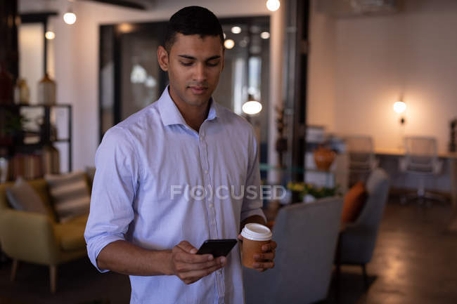 Vista frontale del bel giovane uomo d'affari di razza mista che utilizza il telefono cellulare in ufficio mentre tiene una tazza di caffè contro il soggiorno sullo sfondo — Foto stock