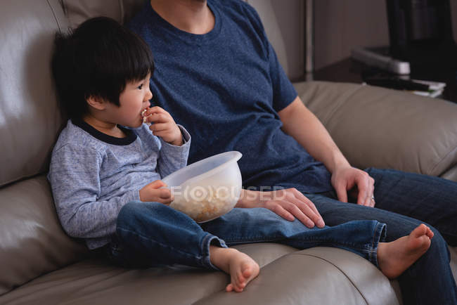 Front view of a happy Asian father and son sitting on sofa and eating popcorn while watching TV at home — Stock Photo