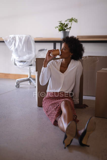 Вид спереду красива бізнес-леді змішаної раси розслабляється під час пиття кави, що сидить на підлозі в сучасному офісі — стокове фото