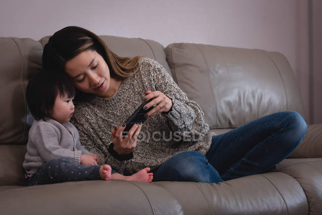 Vista frontal da mãe e da filha asiáticas usando o telefone celular enquanto estão sentados no sofá em casa — Fotografia de Stock