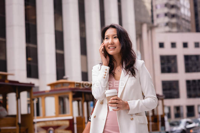 Вид на щасливу азіатській жінці, яка розмовляє на мобільному телефоні, маючи каву на вулиці — стокове фото