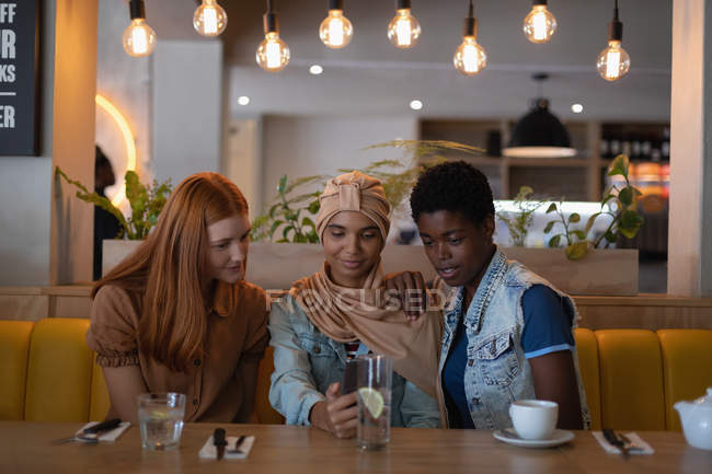 Vista frontal de felices amigas de raza mixta usando teléfono móvil mientras están sentadas en el restaurante - foto de stock
