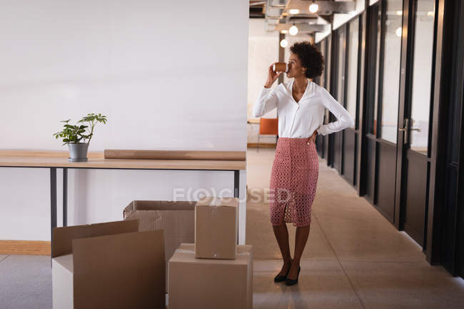 Vista frontale della bella donna d'affari mista che beve caffè in piedi in un ufficio moderno — Foto stock