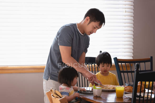 Vista lateral de un padre asiático que sirve el desayuno a sus hijos en la mesa de comedor en la cocina en casa - foto de stock
