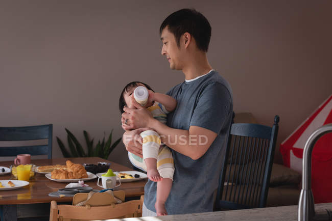 Vista lateral de un padre asiático feliz alimentando a la niña con biberón en la cocina en casa - foto de stock