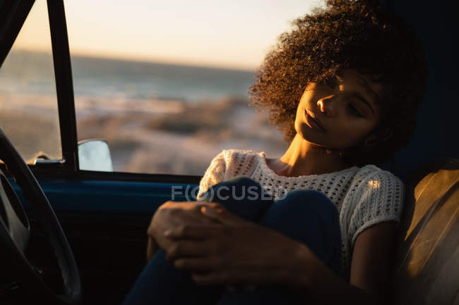 Vista frontal da bela mulher afro-americana pensativa sentada no carro na praia ao pôr do sol — Fotografia de Stock