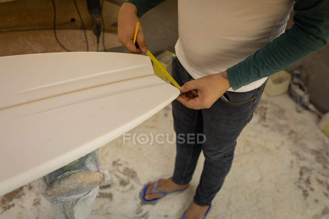 Sección baja del hombre midiendo el final de una tabla de surf en un taller - foto de stock