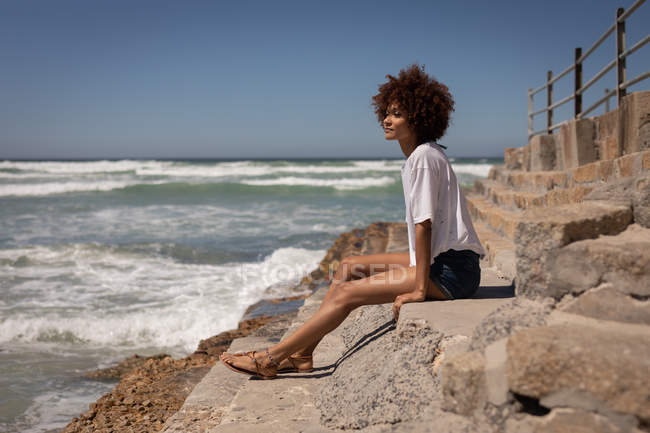 Вид сбоку молодой женщины смешанной расы, расслабляющейся, сидя на скалах на пляже на солнечном берегу — стоковое фото
