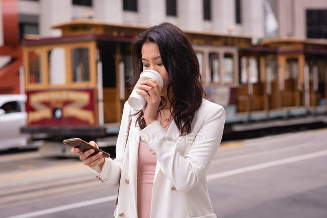 Vista frontal de la mujer asiática reflexiva usando el teléfono móvil mientras toma café en la calle - foto de stock