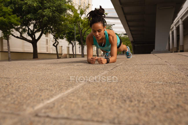 Front view of young Mixed race woman listening music on earphones while doing plank exercise in the city — Stock Photo