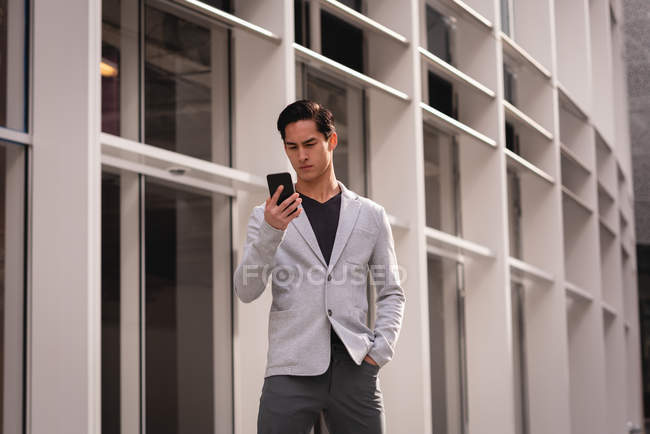 Vista frontal do homem asiático bonito usando telefone celular enquanto está perto do edifício — Fotografia de Stock