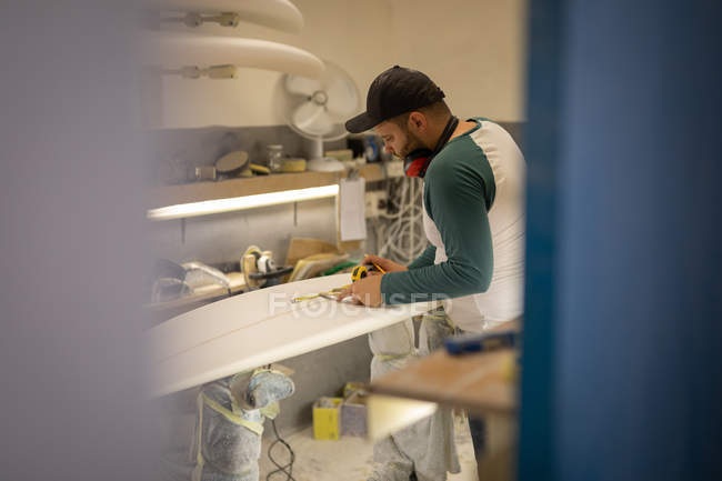 Vista laterale dell'uomo caucasico con paraorecchie che misura tavola da surf con righello e matita in un laboratorio — Foto stock