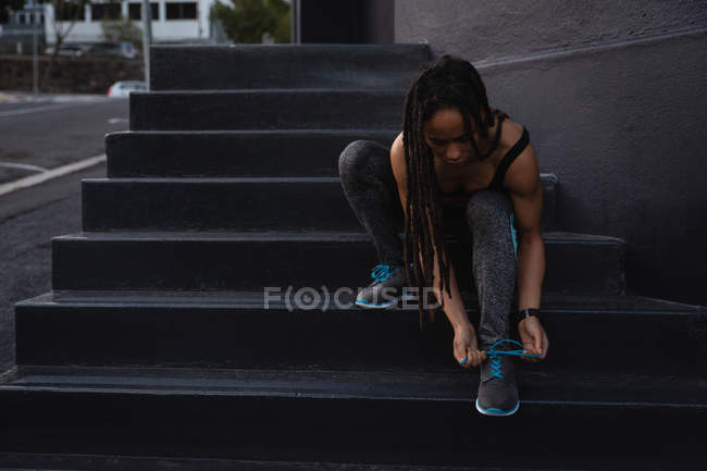 Vista frontale della vestibilità giovane donna di razza mista che si lega i lacci delle scarpe sulle scale della città — Foto stock