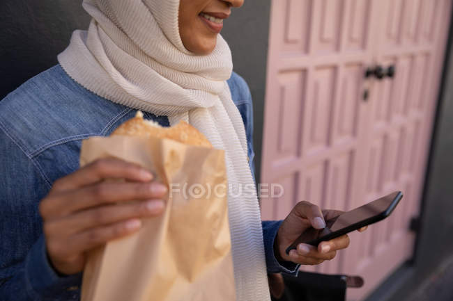 Sección media de una mujer mestiza usando teléfono móvil mientras desayuna en la calle - foto de stock