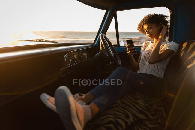 Вид збоку красива молода афро-американської жінки за допомогою мобільного телефону, спираючись в машині на пляжі на заході сонця — стокове фото