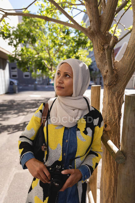 Vue de face d'une jeune femme métissée réfléchie debout dans la rue avec un appareil photo numérique — Photo de stock