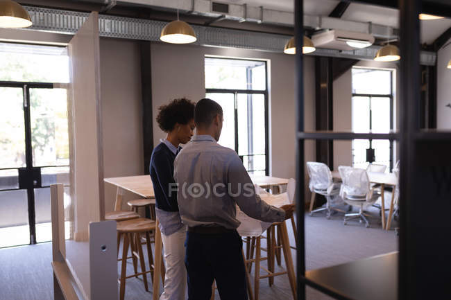 Вид ззаду молодих бізнесменів змішаної раси, які обговорюють синій друк, який тримає чоловік, стоячи в сучасному офісі — стокове фото