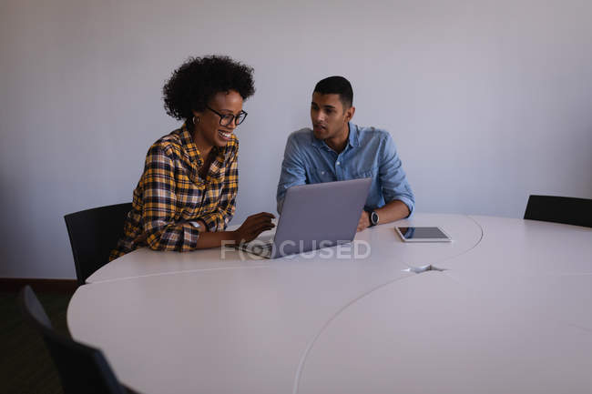 Вид спереду молодих бізнесменів змішаної раси, які обговорюють ноутбук за столом в сучасному офісі — стокове фото