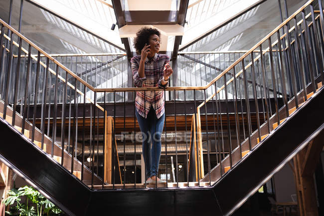 Blick auf die schöne Mischlingshündin, die sich am Geländer lehnt, während sie im modernen Büro am Treppenaufgang im Mobiltelefon spricht — Stockfoto