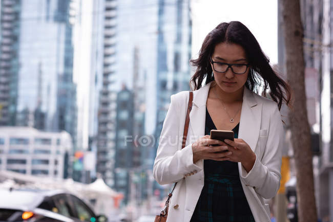 Front view of Asian woman with glasses using her mobile phone while walking in the street on a sunny day — Stock Photo
