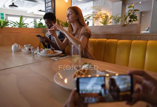 Vue latérale d'amis mixtes prenant des photos de la nourriture du petit déjeuner avec téléphone portable au restaurant — Photo de stock