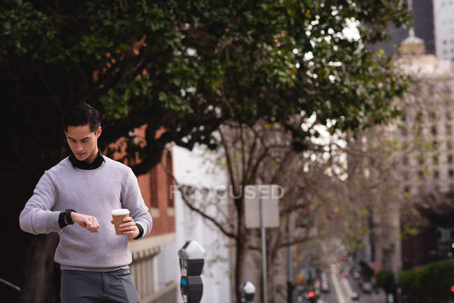 Front view of handsome Asian man using smart watch while having coffee on street — Stock Photo